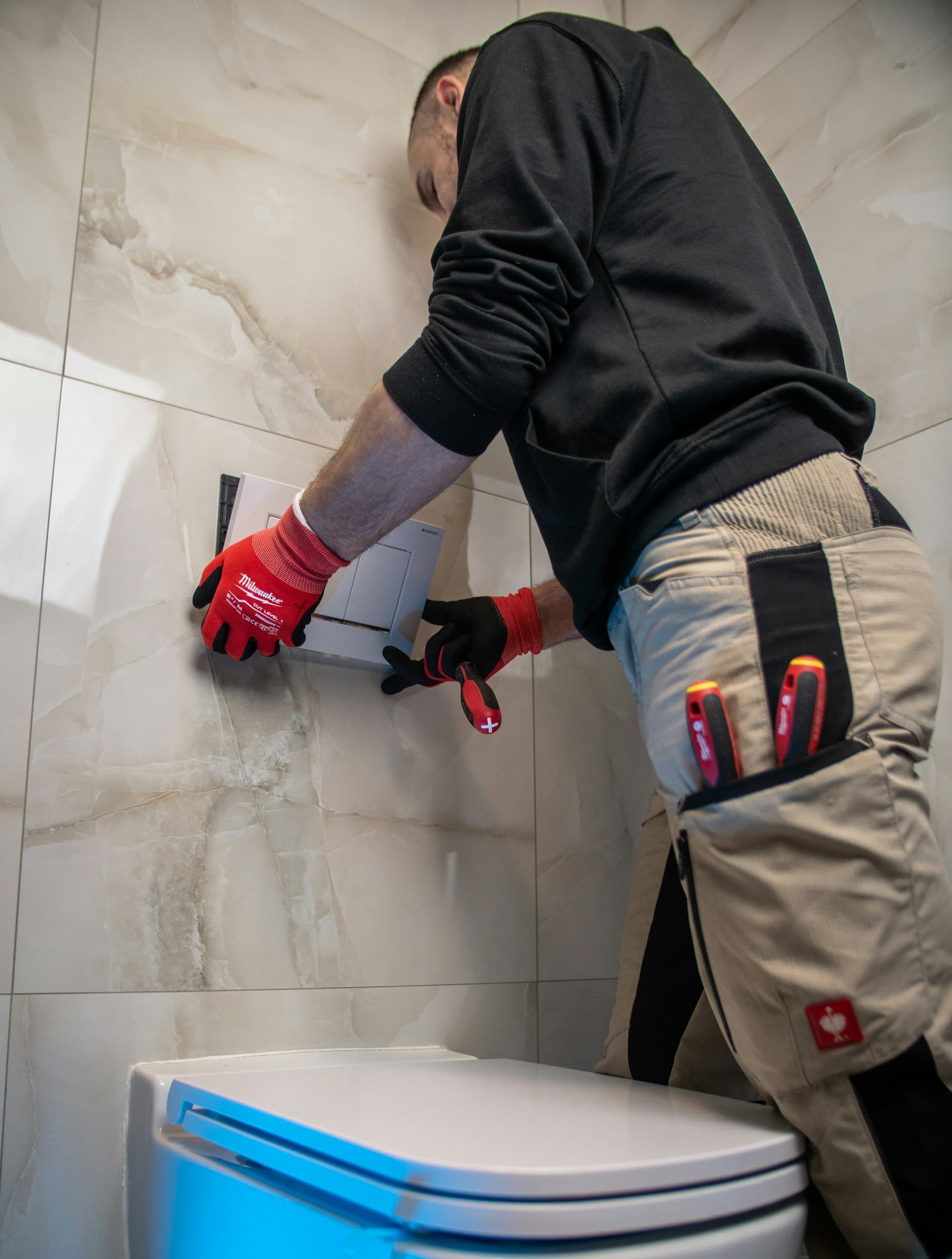 Plumbing engineer in work gloves fitting a flush plate in a tiled bathroom
